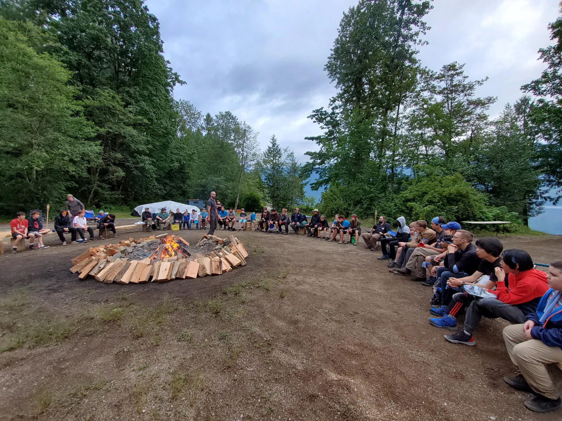 Dusk. Everyone in a circle around the fire pit by the water, mountains behind